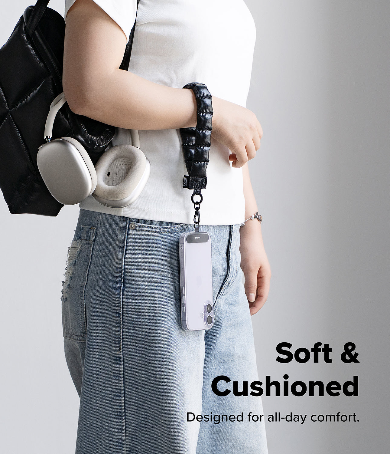 Holder Link Puffer | Hand - Person holding a black backpack with a black puffer hand strap, wearing a white shirt and blue jeans on a gray background.