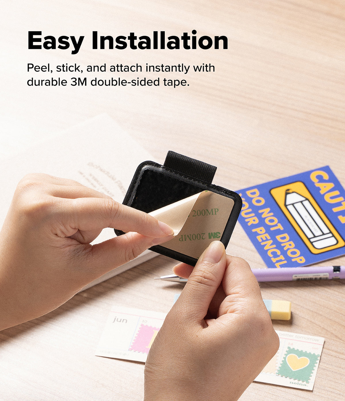 Pen Holder | Name Tag (2P) - A close-up of hands demonstrating the easy installation of a small black adhesive card or label with double-sided tape on a light wood desk. Text at the top reads 'Easy Installation' and lists peel, stick, and attach instructions.