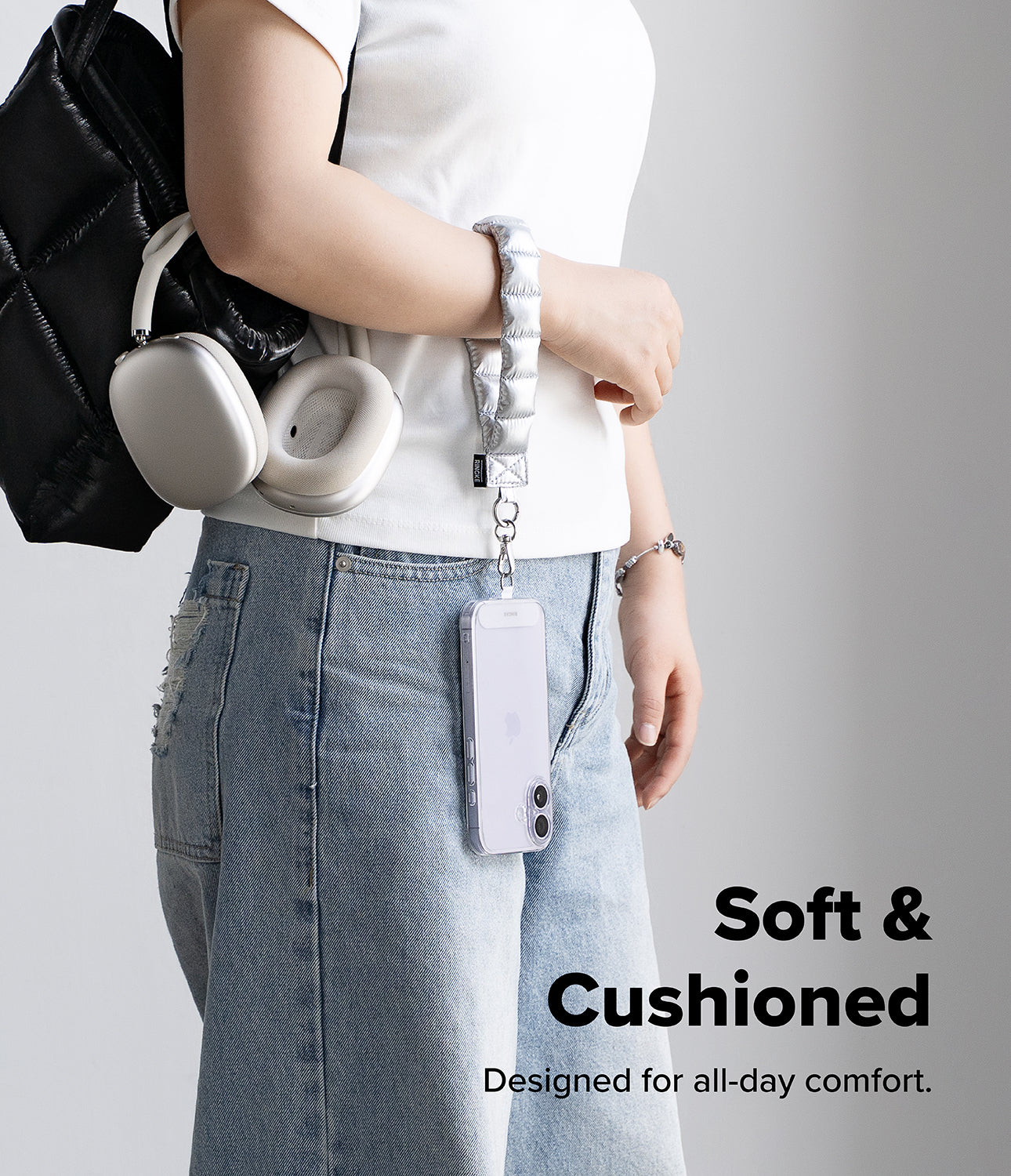 Holder Link Puffer | Hand - Person holding a black backpack with a silver puffer hand strap, wearing a white shirt and blue jeans on a gray background.