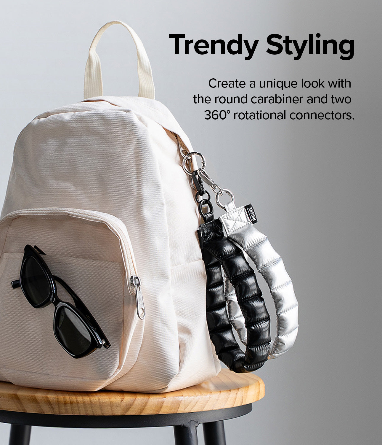 Holder Link Puffer | Hand - Cream backpack on a wooden stool with black and silver puffer straps attached by a carabiner.