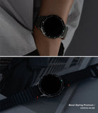 Galaxy Watch 9 / 8 44mm | Bezel Styling Premium - A smartwatch with a black screen and a dark gray textured strap is shown worn on a person's wrist in the top half, and resting on a dark surface in the bottom half. The watch has a black bezel with small colored markers and a small orange accent on the strap.