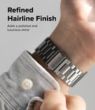 Galaxy Watch 9 / 8 | Metal One Air - A close-up of a silver stainless steel link watch band worn on a wrist, with a light denim shirt cuff visible. Text on the left reads 'Refined Hairline Finish' and 'Adds a polished and luxurious shine'.