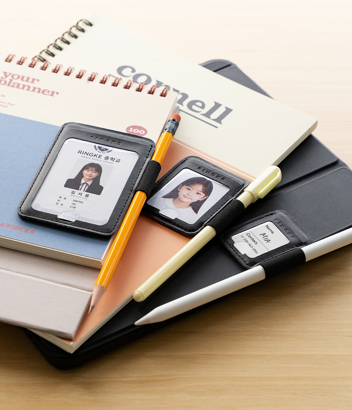Pen Holder | Name Tag (2P) - A stack of stationery items on a light wood surface, including notebooks, a pencil, a pen, and multiple black ID badge holder cases with sample photo cards.