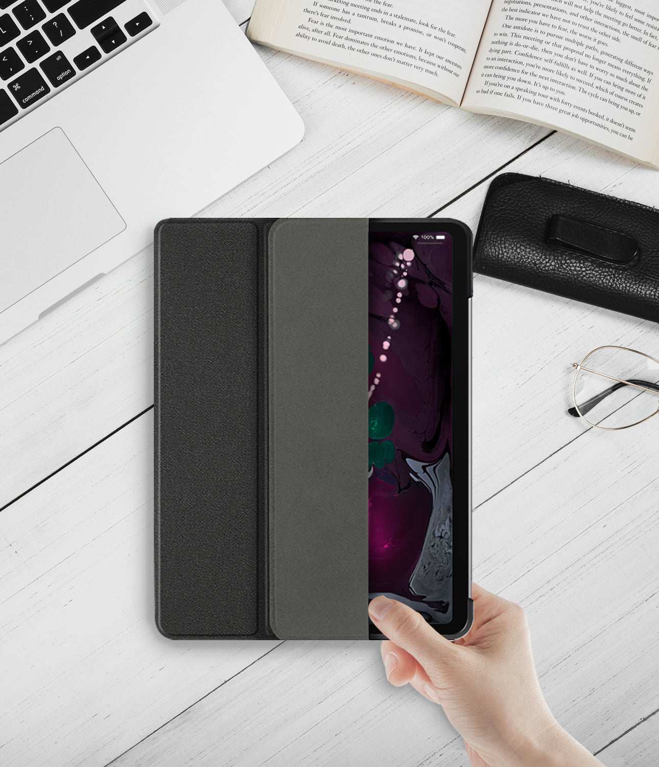 Smart Cover Apple Pencil Stand | iPad - A black folio-style tablet case with a built-in kickstand, shown open and held by a hand on a white wooden desk. A laptop, open book, glasses, and a black leather accessory case are visible around it.