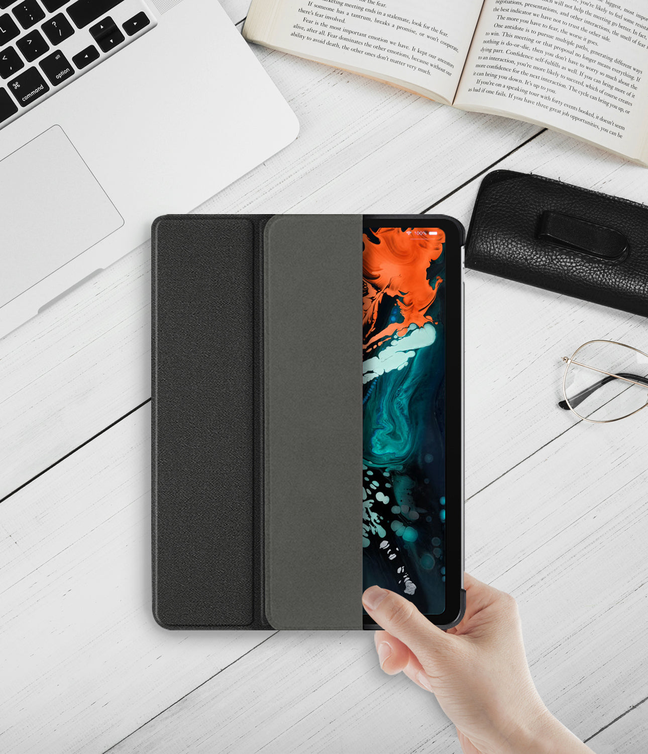 Smart Cover Apple Pencil Stand | iPad - A black folio-style smartphone case with a built-in screen protector is shown being held by a hand on a white wooden desk, surrounded by lifestyle props including a laptop, an open book, glasses, and a black leather pouch.
