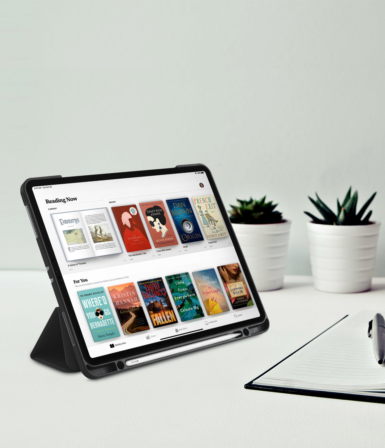 Smart Cover Apple Pencil Stand | iPad - A tablet in a black protective case with a built-in kickstand, shown on a white desk with a notebook and pen, and two small potted succulents in the background.