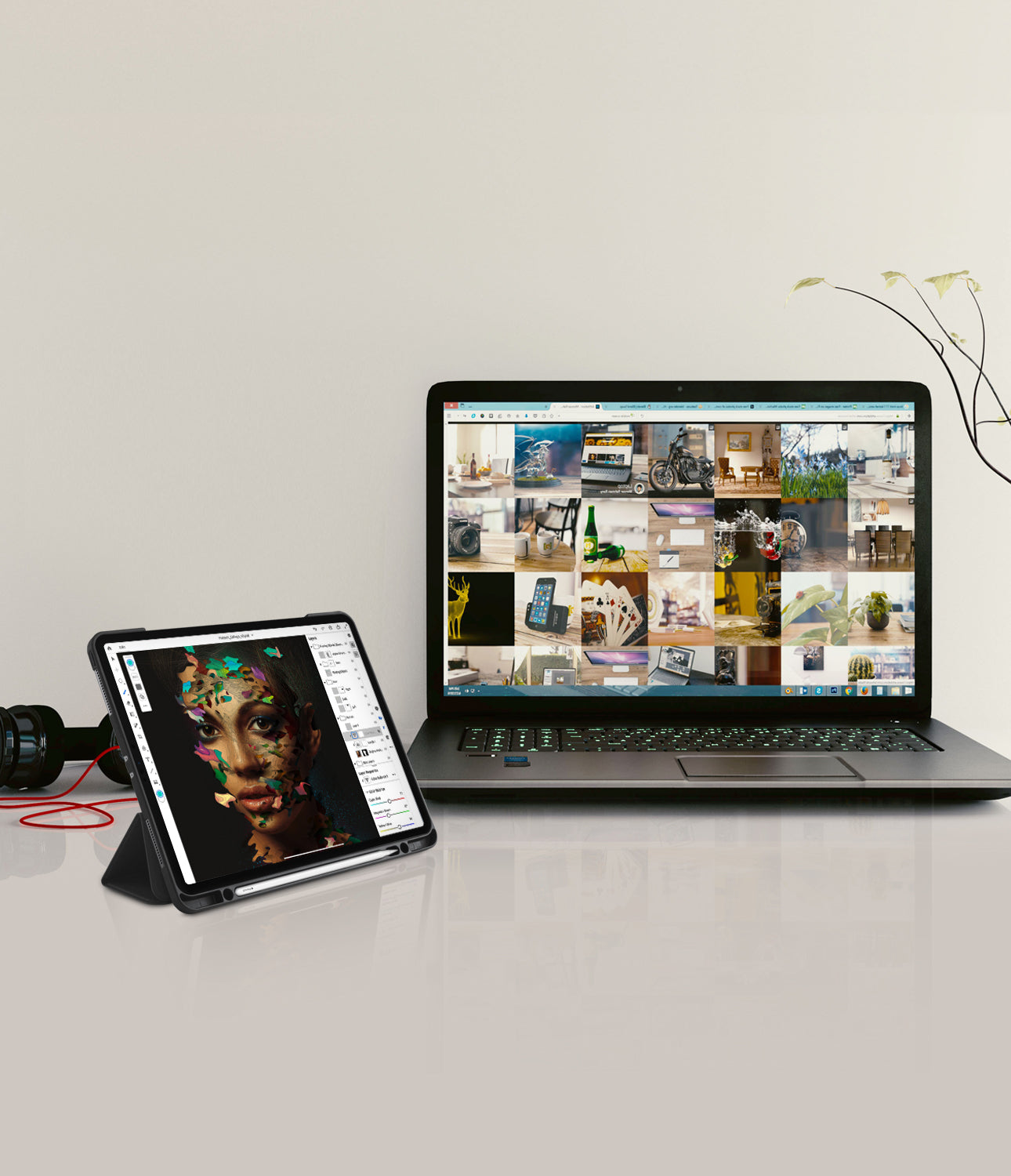 Smart Cover Apple Pencil Stand | iPad - A black tablet in a fold-out stand displaying a colorful portrait, placed beside an open silver laptop showing a collage of interior design images on a light gray surface against a beige wall.