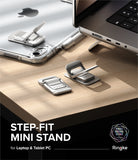 Step-Fit Mini Stand - Two silver metallic step-fit mini laptop and tablet stands shown on a wooden desk, with a laptop and tablet partially visible in a styled workspace scene. Text at the bottom reads 'STEP-FIT MINI STAND for Laptop & Tablet PC' and shows the Ringke brand logo.