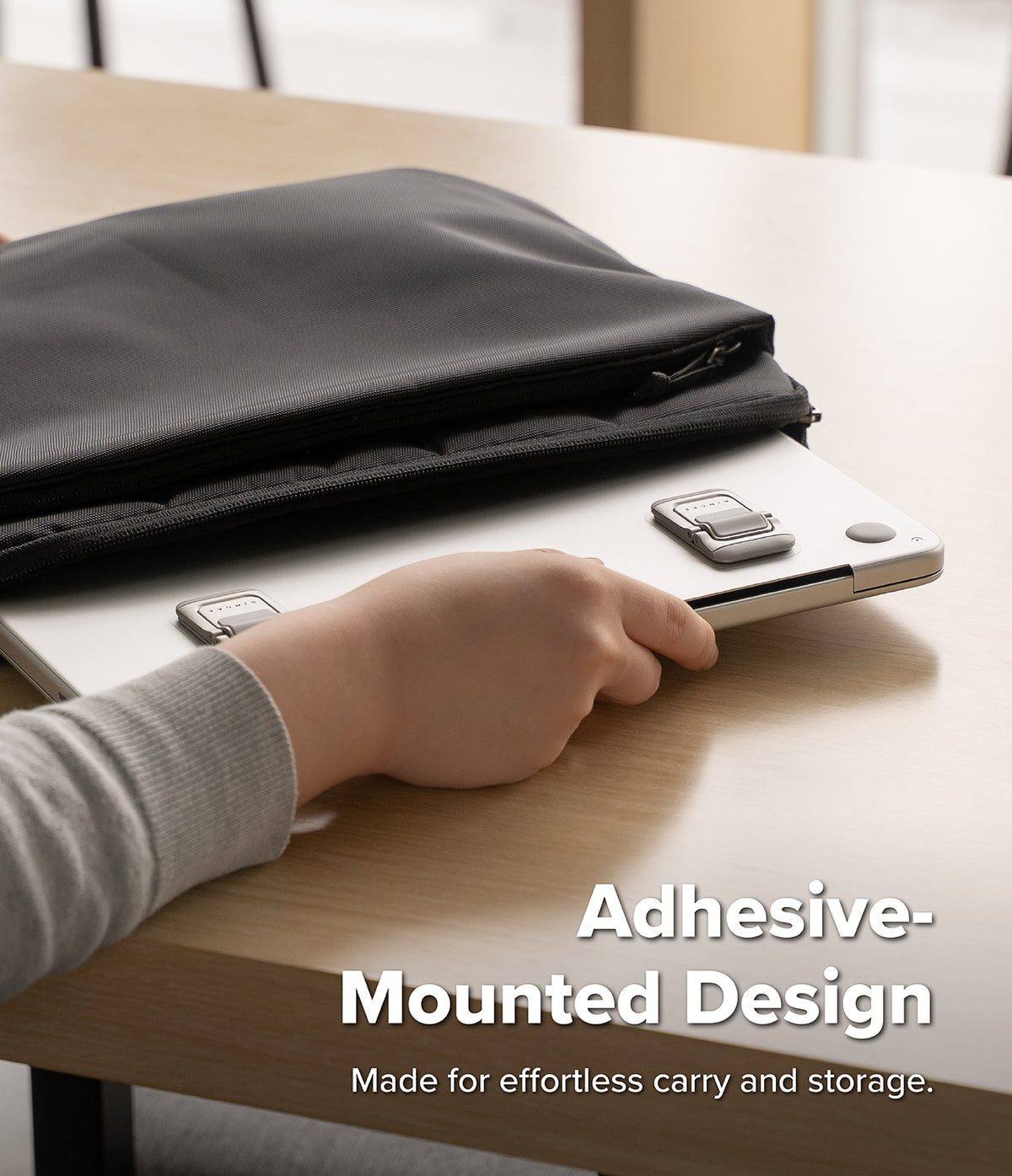 Step-Fit Mini Stand - A person's hand is shown attaching a silver laptop to a black adhesive laptop sleeve on a light wood desk. The text overlay reads 'Adhesive-Mounted Design' and 'Made for effortless carry and storage.